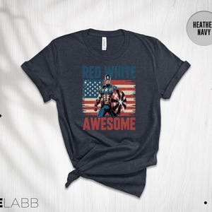May include: A heather navy short-sleeve t-shirt featuring a graphic of Captain America standing in front of a distressed American flag. The text "RED WHITE" appears in blue above the character, and "AWESOME" is printed in red below the graphic.