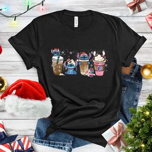 May include: Black t-shirt with a graphic of four Stitch characters holding different holiday drinks. The characters are wearing Santa hats and holding cups of coffee, hot chocolate, and eggnog. The background is a white snowflake pattern.