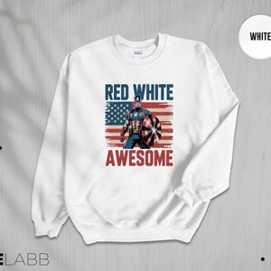 May include: A white crewneck sweatshirt featuring a graphic print of a superhero holding a shield in front of a distressed American flag. The text RED WHITE is printed in blue above the figure, and the word AWESOME is printed in red below.