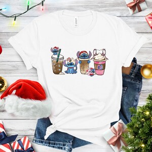 May include: White t-shirt with a graphic of four Stitch characters dressed as different holiday drinks. The characters are wearing Santa hats and holding cups of coffee, hot chocolate, and iced coffee.