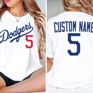 May include: A white t-shirt featuring the blue script text Dodgers with a red number 5 on the front. The back of the t-shirt displays the text CUSTOM NAME in blue above a large blue number 5. The shirt has short sleeves and a crew neckline.