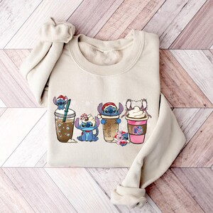 May include: A beige sweatshirt with a graphic print of four Stitch characters holding different coffee drinks. The characters are wearing Santa hats and holding cups with whipped cream and sprinkles.