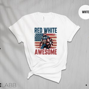 May include: A white short-sleeved t-shirt featuring a graphic of a superhero holding a shield in front of a distressed American flag. The text RED WHITE is printed in blue above the figure, and the word AWESOME is printed in red below the figure.