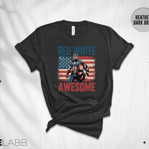 May include: A heather dark grey t-shirt featuring a graphic of a superhero holding a shield in front of a distressed American flag. The text "RED WHITE" appears in blue above the figure, and the word "AWESOME" is printed in red below the graphic.
