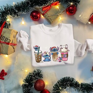 May include: White sweatshirt with a Christmas-themed design featuring Stitch characters holding cups of coffee and cocoa.