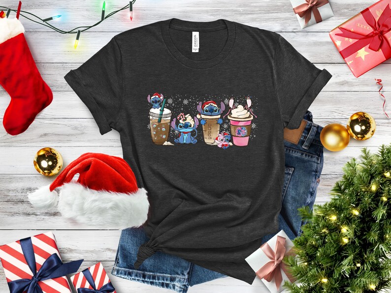 May include: A dark gray t-shirt with a Christmas-themed design featuring four Stitch characters holding different holiday drinks. The characters are wearing Santa hats and the drinks are decorated with snowflakes.