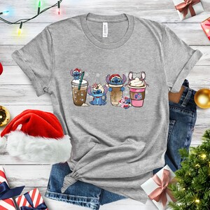 May include: A gray t-shirt with a Christmas-themed design featuring four Stitch characters holding different holiday-themed drinks. The characters are wearing Santa hats and scarves. The background is a white snowflake pattern.