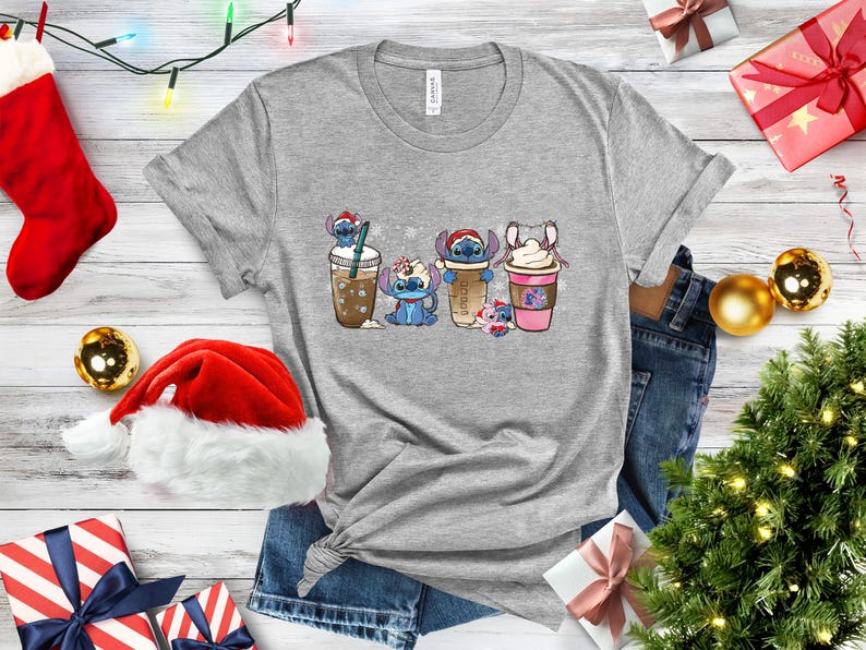 May include: A gray t-shirt with a Christmas-themed design featuring four Stitch characters holding different holiday-themed drinks. The characters are wearing Santa hats and scarves. The background is a white snowflake pattern.