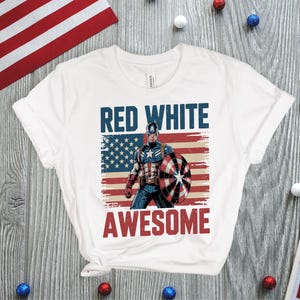 May include: A white short-sleeve t-shirt featuring a graphic of Captain America holding a shield in front of a distressed American flag. The text "RED WHITE" appears in blue above the character, and the word "AWESOME" is printed in red below.