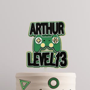 May include: A green and black cake topper featuring a video game controller and the text "Arthur Level 13". The cake topper is on a white cake decorated with green and gold geometric shapes.
