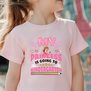 May include: A pink t-shirt with a graphic design that says "My Princess is going to Kindergarten" with a cartoon princess, stars, hearts, and a pencil.