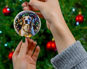 May include: A round white Christmas bauble featuring a photograph of a dog wearing a string of Christmas lights. The dog is a brown and white hound with a black nose and brown eyes. The bauble is suspended from a silver string.