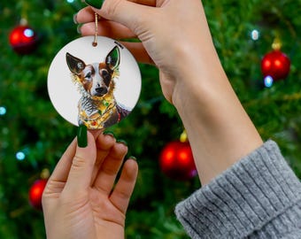May include: A round white ceramic ornament with a colorful image of a dog wearing a festive garland. The dog has brown, white, and black fur.