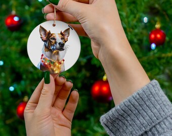 May include: A circular ceramic Christmas decoration depicting a dog with brown and white fur, adorned with a festive, illuminated collar. The ornament is held against a Christmas tree with red baubles and twinkling lights.