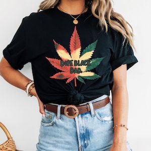 May include: Black t-shirt featuring a colorful cannabis leaf design with the text "DOPE BLACK DAD." The leaf is colored with red, yellow, and green hues. The shirt is paired with denim shorts and a brown belt.
