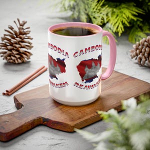 May include: White ceramic mug with a pink handle and rim, featuring a map of Cambodia with the Cambodian flag and the words "Cambodia, Be Awaken". The mug is filled with a dark liquid and sits on a wooden chopping board.