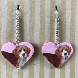 May include: A pair of dangle earrings featuring a pink heart-shaped charm with a beagle dog image. The earrings have a silver chain.