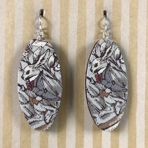 Puede incluir: Un par de pendientes ovalados con una ilustración de conejo blanco sobre fondo gris. El conejo lleva una pajarita y tiene una expresión de sorpresa.