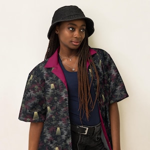 May include: A dark charcoal bucket hat and a short-sleeved button-down shirt featuring a pattern of gray alien heads, flying saucers with yellow light beams, and cacti over a dark camouflage background with a solid magenta inner collar lining.