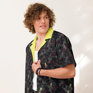 May include: A short-sleeved button-down shirt featuring a dark camouflage pattern with alien, UFO, and cactus motifs. The shirt has a bright lime green collar and lapels, worn over a plain white undershirt, showcasing a casual and distinct design.
