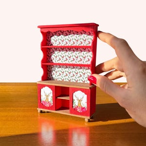 May include: A miniature red wooden cabinet with shelves and a lower cabinet. The shelves and back of the cabinet are decorated with a floral pattern. The lower cabinet doors feature a deer illustration. The cabinet is on a wooden surface.