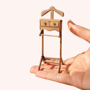 Puede incluir: Miniatura de madera con perchero, dos cajones pequeños y una barra inferior. El soporte es de color marrón claro y está sostenido en la mano de una persona. El soporte es probablemente para ropa de muñecas o una colección en miniatura.