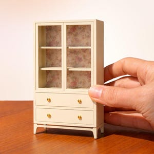 Könnte beinhalten: Ein cremefarbener Miniaturschrank mit Glastüren und zwei Schubladen. Der Innenraum hat Regale und eine florale Tapete. Goldfarbene Knöpfe zieren die Schubladen. Der Schrank ist für ein Puppenhaus oder eine Miniaturumgebung.