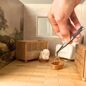 Hand using tweezers to place a tiny woven basket inside a 1:12 scale dollhouse nursery scene with miniature furniture, crib, and dresser. The handmade miniature basket is displayed in a dollhouse interior, showing the finished DIY dollhouse basket for miniature decor and dollhouse accessories.