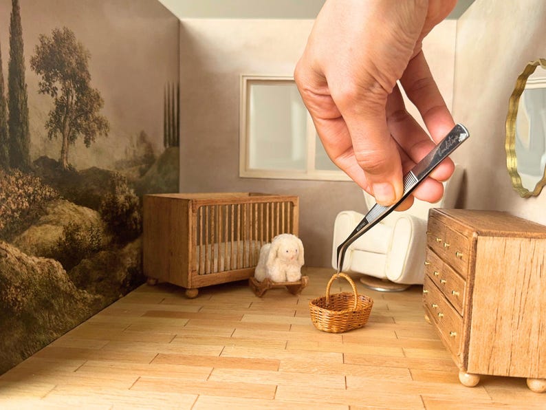 Hand using tweezers to place a tiny woven basket inside a 1:12 scale dollhouse nursery scene with miniature furniture, crib, and dresser. The handmade miniature basket is displayed in a dollhouse interior, showing the finished DIY dollhouse basket for miniature decor and dollhouse accessories.