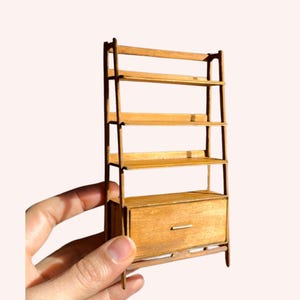 Puede incluir: Una estantería de madera en miniatura con cinco estantes y un cajón. La estantería está hecha de madera de color claro y tiene un diseño simple y moderno. El cajón tiene un pequeño tirador redondo. La estantería está sujeta por una mano.