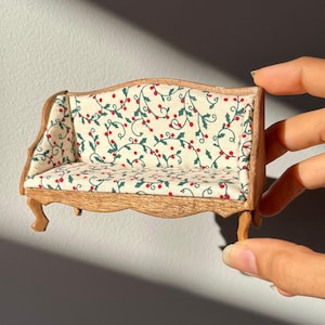 May include: Miniature wooden sofa with a cream-colored fabric featuring a pattern of green leaves and red berries. The sofa has curved arms and legs, and is designed for a dollhouse or similar setting.