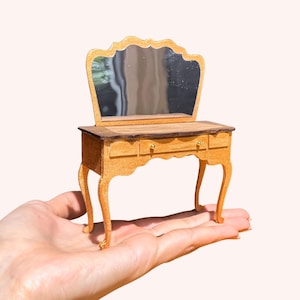 May include: Miniature wooden dressing table with a mirror. The dressing table has two drawers and curved legs. The mirror has a decorative top. The wood is light brown with a darker brown top. The vanity is held in a hand.