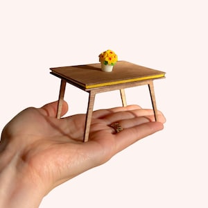 May include: Miniature wooden table with a yellow accent and a small vase of yellow flowers. The table is held in a hand, showcasing its small size. The table measures approximately 2.5 inches square.