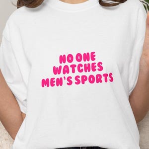 May include: White t-shirt with the text "NO ONE WATCHES MEN'S SPORTS" in bright pink, bubble letters. The shirt is a simple, casual style.
