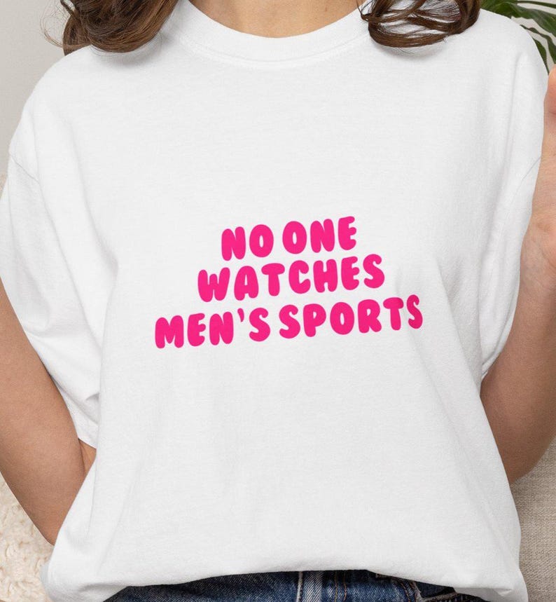 May include: White t-shirt with the text "NO ONE WATCHES MEN'S SPORTS" in bright pink, bubble letters. The shirt is a simple, casual style.