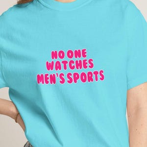 Puede incluir: Camiseta azul claro con el texto rosa "NO ONE WATCHES MEN'S SPORTS" en letras de burbujas. La camiseta de manga corta est&aacute; hecha de un tejido suave.