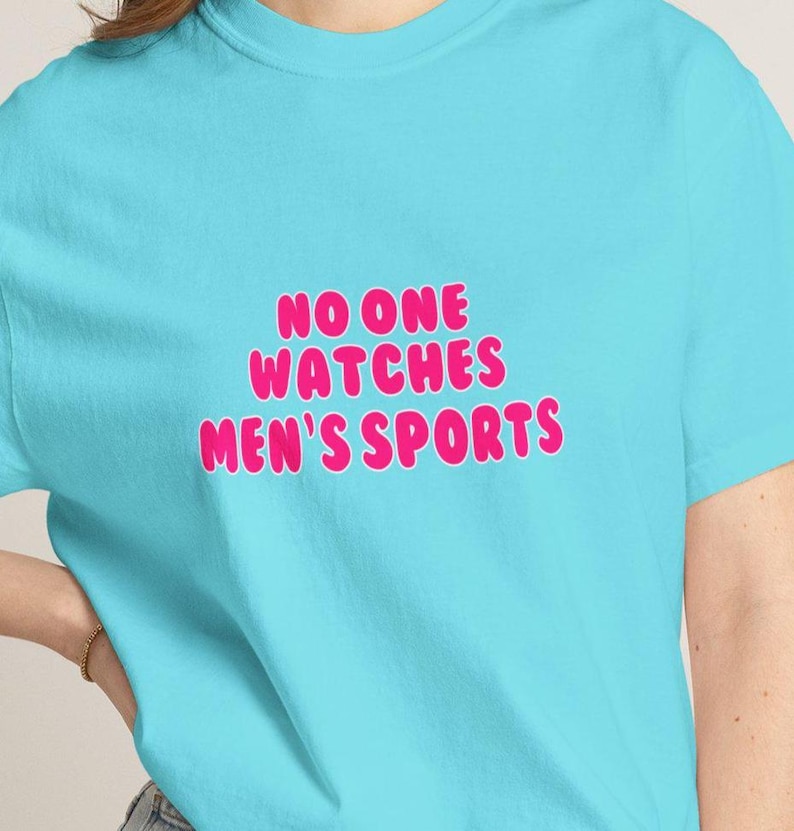 Puede incluir: Camiseta azul claro con el texto rosa "NO ONE WATCHES MEN'S SPORTS" en letras de burbujas. La camiseta de manga corta est&aacute; hecha de un tejido suave.