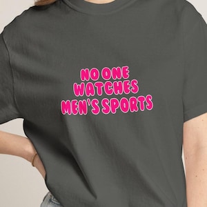 May include: Dark gray t-shirt with the text "NO ONE WATCHES MEN'S SPORTS" in pink bubble letters. The text has a white outline. The shirt is a casual wear item.