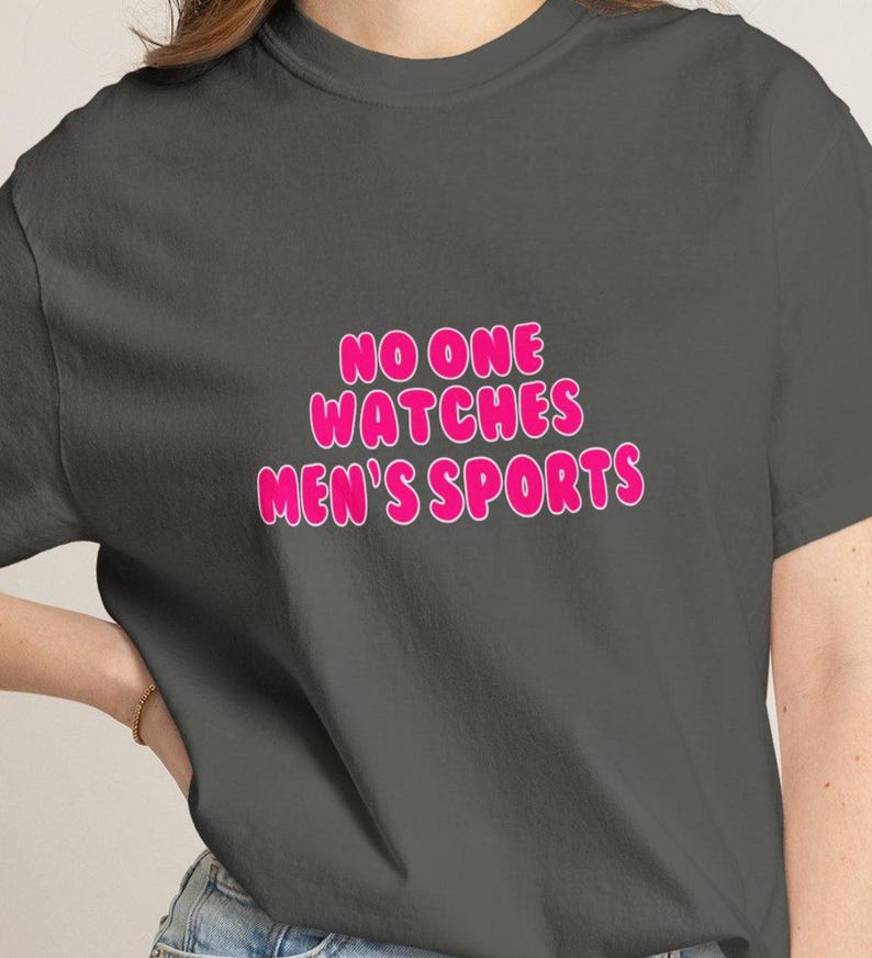 May include: Dark gray t-shirt with the text "NO ONE WATCHES MEN'S SPORTS" in pink bubble letters. The text has a white outline. The shirt is a casual wear item.