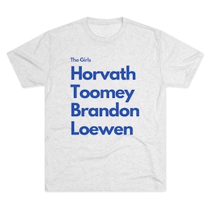 May include: A light gray t-shirt with the words "The Girls" in small blue text at the top. Below, in large blue text, are the names Horvath, Toomey, Brandon, and Loewen. The shirt is made of a soft, comfortable material.