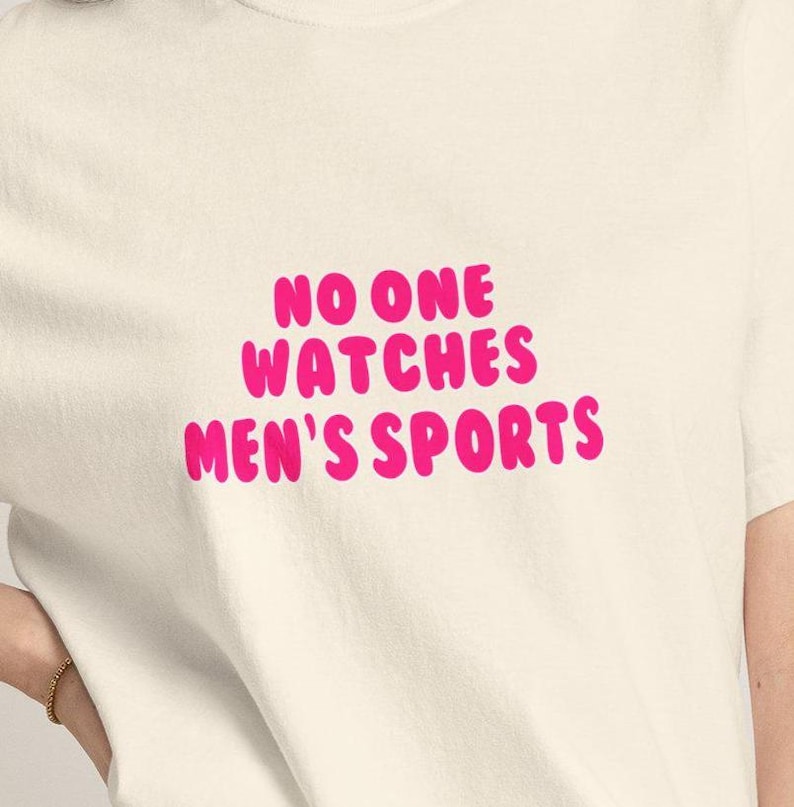 K&ouml;nnte beinhalten: Cremefarbenes T-Shirt mit dem Text "NO ONE WATCHES MEN'S SPORTS" in leuchtend pinkfarbenen, fetten Buchstaben. Das Shirt besteht aus einem weichen Material und ist f&uuml;r den t&auml;glichen Gebrauch geeignet.