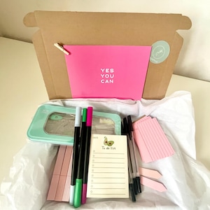 May include: A light blue metal box with a clear lid, filled with pink, green, and black pens, a to-do list notepad, and pink paper clips. The box is in a brown cardboard box with a pink card that says "YES YOU CAN".