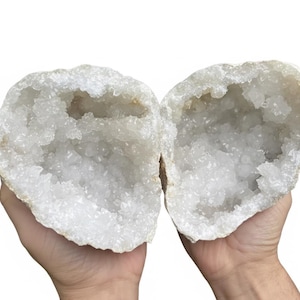 May include: Two halves of a geode, revealing a sparkling interior of white quartz crystals. The rough, beige exterior contrasts with the crystalline interior. The geode is held open, showcasing the intricate crystal formations. A natural, decorative item.