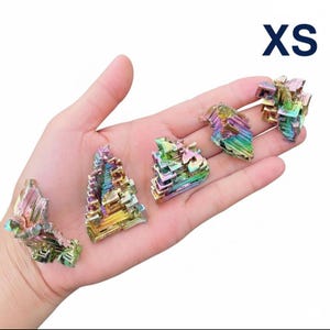 May include: Five small, iridescent bismuth crystals in various shapes are displayed in a hand. The crystals exhibit a range of colors, including blues, greens, yellows, and purples. The text "XS" is in the upper right corner.