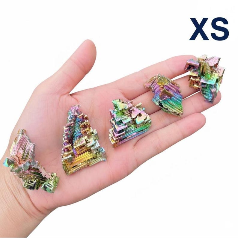 May include: Five small, iridescent bismuth crystals in various shapes are displayed in a hand. The crystals exhibit a range of colors, including blues, greens, yellows, and purples. The text "XS" is in the upper right corner.