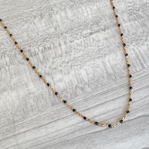 May include: Gold-toned necklace with small, black beads evenly spaced along the chain. The necklace is displayed against a light gray wooden surface, creating a contrast between the jewelry and the background.