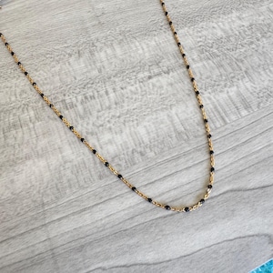 May include: A delicate gold-colored necklace featuring small, evenly spaced black beads. The necklace has a fine chain and a simple, elegant design. The beads are a deep black, contrasting with the gold chain. The necklace is displayed on a light gray wooden surface.