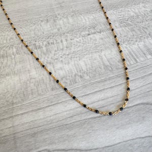 May include: A delicate gold-tone necklace featuring small, evenly spaced black beads. The necklace has a fine chain and a simple, elegant design. The beads are a deep black color, creating a contrast with the gold chain.