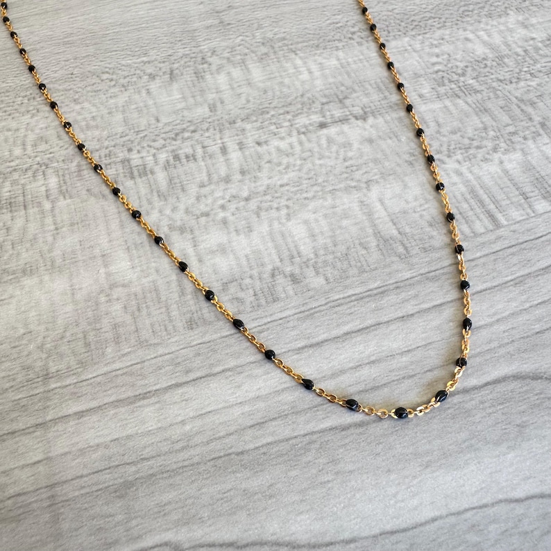May include: A delicate gold-tone necklace featuring small, evenly spaced black beads. The necklace has a fine chain and a simple, elegant design. The beads are a deep black color, creating a contrast with the gold chain.