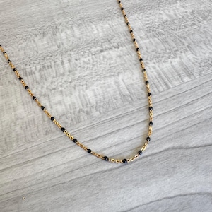 May include: A delicate gold-toned necklace featuring small, evenly spaced black beads. The necklace has a fine chain and a simple, elegant design. The beads are a deep, glossy black, contrasting with the warm gold color of the chain.